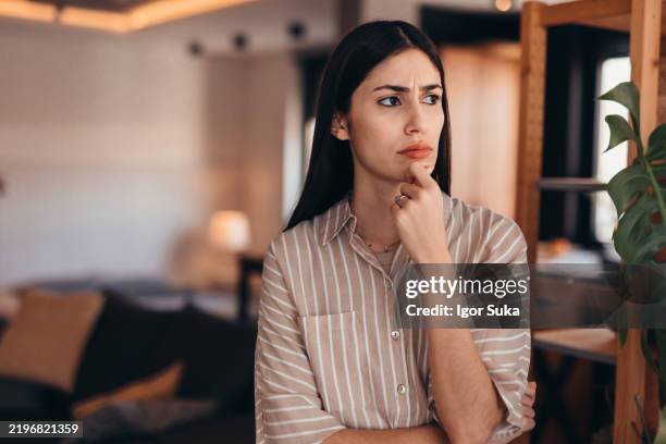 young woman looking pensive while standing in living room - verward stockfoto's en -beelden