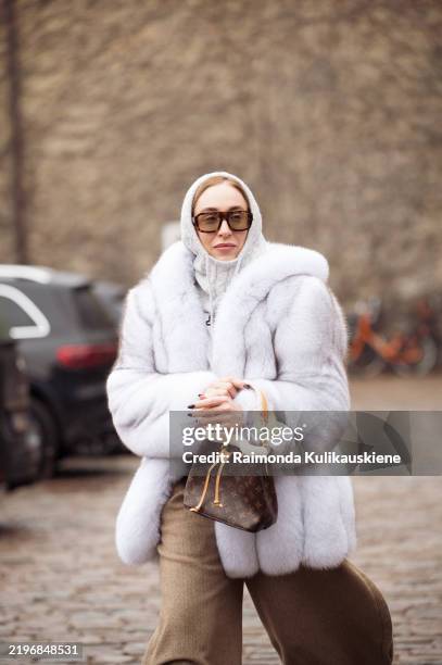 Sonia Lyson wears beige wide pants, grey balaclava, pale blue fur coat, brown bag, sunglasses and white and grey sneakers outside the Munthe fashion...