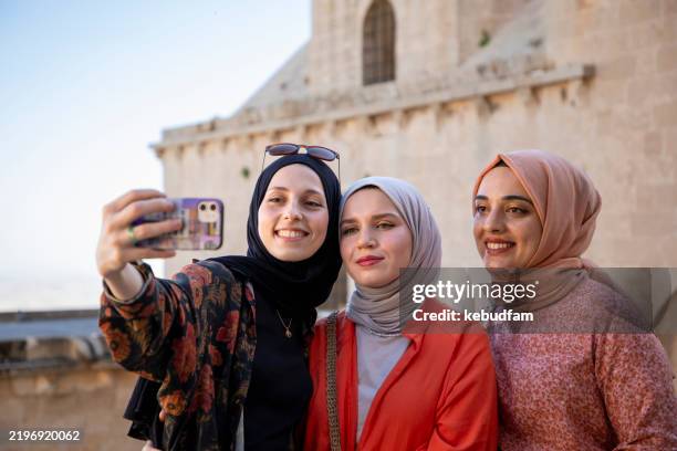 three young women wearing hijabs are taking a selfie in mardin, turkey. they are smiling, dressed stylishly, and standing in front of an old stone building with arches and domes. the sky is clear and blue. - mardin stock pictures, royalty-free photos & images