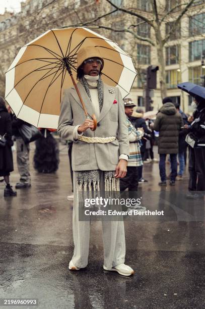 Guest wears a hat, a gray oversized blazer jacket, suit pants, sneakers, holds an umbrella, outside Hermes, during the Menswear Fall Winter 2025/2026...
