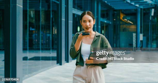 young asian businesswoman records voice note on her smartphone while holding digital tablet while walking outside office building. remotely work and sustainable business outdoor. - conference phone stock pictures, royalty-free photos & images