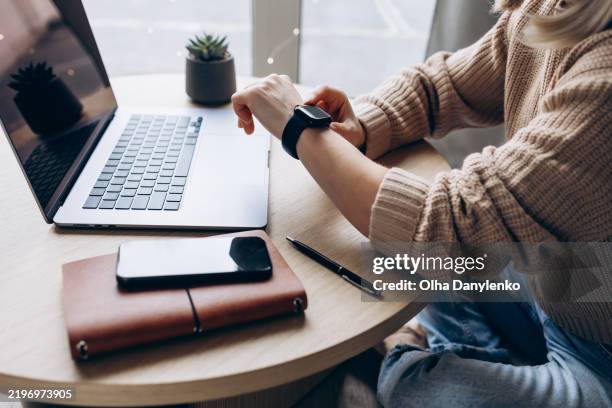 woman checking smartwatch while working on laptop at cozy wooden desk with notebook, smartphone, and pen in daylight. successful female entrepreneur in business. remote-working. time management and productivity. business on the go concept - checking the time stock pictures, royalty-free photos & images