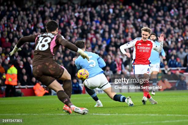 Martin Odegaard of Arsenal scores his team's first goal whilst under pressure from John Stones of Manchester City as Stefan Ortega of Manchester City...