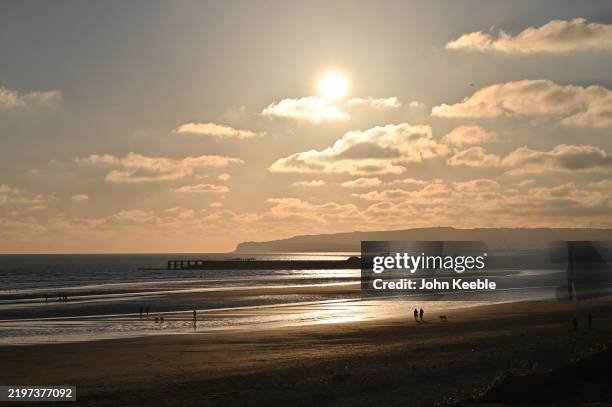 Two people walk their dog along Camber Sands Beach at sunset on February 1, 2025 in Camber, England.