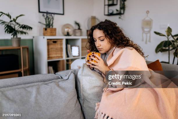 eine frau liegt zusammengekauert auf ihrem sofa, eingekuschelt in eine große decke um ihre schultern, während sie eine tasse heißen tee in der hand hält, um sich aufzuwärmen und sich besser zu fühlen, während sie an einer erkältung oder grippe leid - winterdepression stock-fotos und bilder