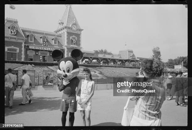 Costumed Mickey Mouse performer poses with a girl as a woman takes their photo in front of the Santa Fe & Disneyland Railroad station at Disneyland,...