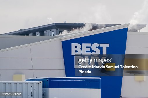 A close-up shot of the Best Buy logo on a building, with a blue and yellow design. Smoke or steam appears to be rising from behind the building. A close-up shot of the Best Buy logo on a building, with a blue and yellow design. Smoke or steam appears to be rising from behind the building.