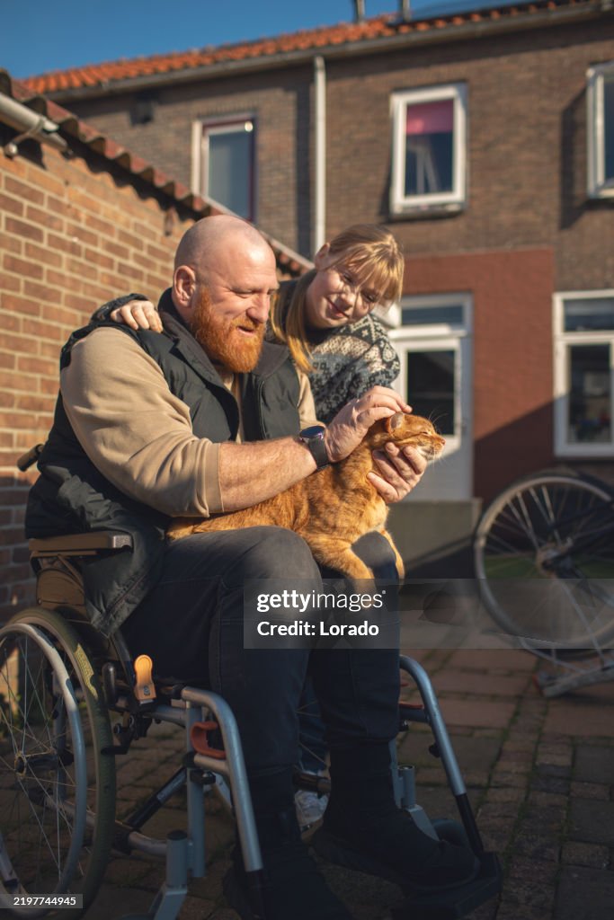 A disabled redhead father and his beautiful blonde daughter with a therapy cat in the garden