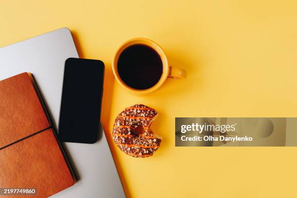 minimalist workspace setup featuring a leather notebook, smartphone, laptop, a yellow coffee cup, and a half-eaten chocolate-glazed donut on a vibrant yellow background - phone flat lay stock pictures, royalty-free photos & images
