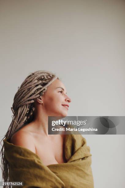a 30-39-year-old woman, looking towards the window, hugs herself, wrapped in a blanket, a light wall behind her, light dreadlocks. cleavage and exposed neck and shoulder - dekolleté stock-fotos und bilder
