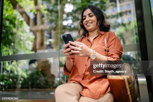 woman enjoying her time while using a smartphone in a park in sydney, australia during a sunny day - bus bench stock pictures, royalty-free photos & images