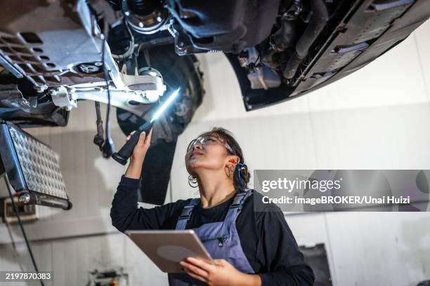 young female mechanic using flashlight and digital tablet, inspecting underside of car raised on lift in auto repair shop - car underside stock pictures, royalty-free photos & images