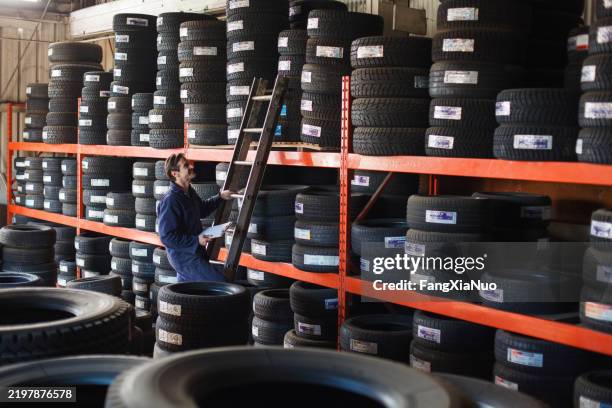 mecánico sosteniendo una llanta de automóvil nueva mirando hacia arriba a la estantería del inventario de llantas para hacer una reparación de vehículo en un garaje de automóviles - neumático fotografías e imágenes de stock