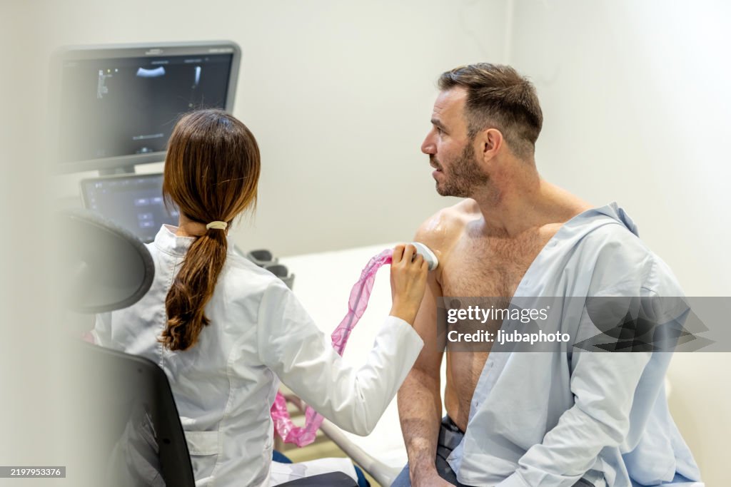 Mid adult Caucasian male patient undergoing shoulder ultrasound examination by diagnostic female doctor.