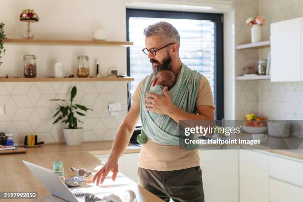 father working from home office while carrying sleeping newborn in baby wrap. man holding a baby and working on laptop in kitchen. - modern manhood stock pictures, royalty-free photos & images