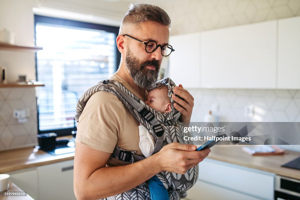 Father scrolling on smartphone while newborn child sleeping in baby carrier. Concept of paternity leave and dad as caregiver for baby.