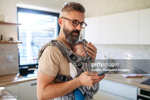 father scrolling on smartphone while newborn child sleeping in baby carrier. concept of paternity leave and dad as caregiver for baby. - mascolinità foto e immagini stock