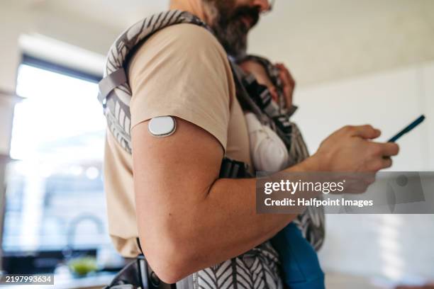 diabetic man with continuous glucose monitor on arm carry his newborn baby in baby carrier. father checking cgm app on smartphone. - continuous glucose monitor stock pictures, royalty-free photos & images