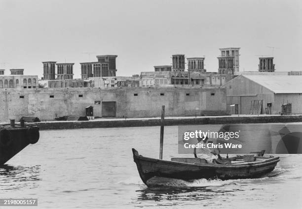 View of a wooden boat on Dubai Creek with traditional buildings featuring wind towers shown in the background, United Arab Emirates, circa May 1967.