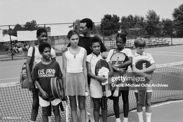 American tennis player Arthur Ashe, wearing a black t-shirt with lighter detail on the right shoulder, poses with children holding their racquet...