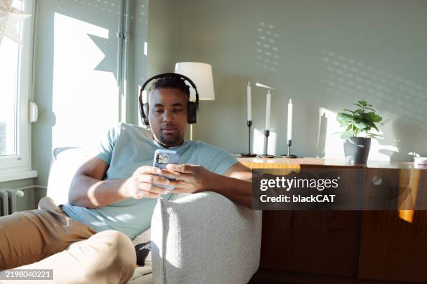 african american man relaxing on a couch with headphones and using a smartphone - virtual assistant stock pictures, royalty-free photos & images