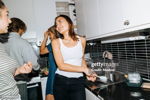 smiling young female student doing dishes while talking with roommate standing in kitchen of college dorm room - flatmate stock pictures, royalty-free photos & images