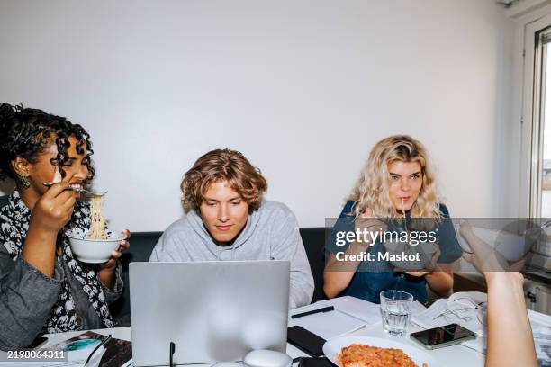 male student using laptop while female roommates eating noodles sitting at desk in college dorm - mund benutzen stock-fotos und bilder