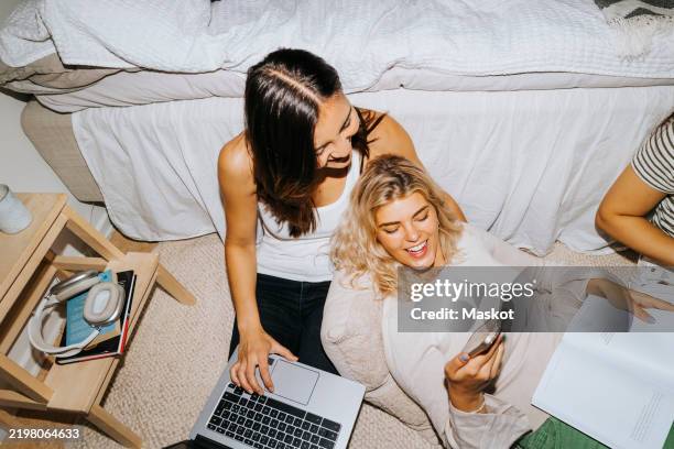 high angle view of smiling woman sharing smart phone with female friend sitting by bed in college dorm - studentenflats stockfoto's en -beelden