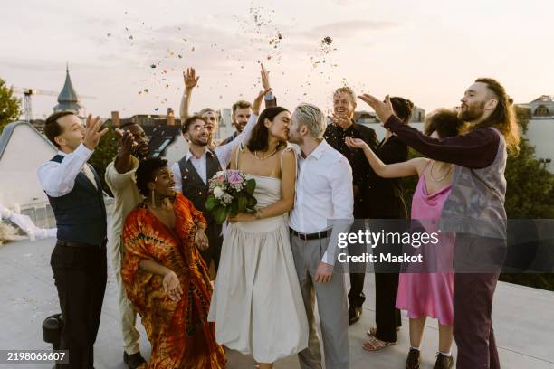 happy wedding guests throwing confetti on newlywed couple kissing on rooftop during wedding celebration - noivo papel em casamento imagens e fotografias de stock