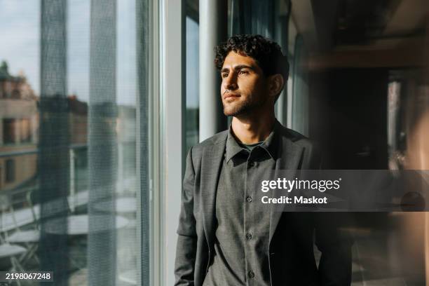thoughtful young male entrepreneur looking through glass window at office - guardare fuori dalla finestra foto e immagini stock