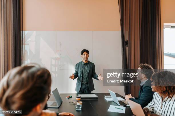 young male entrepreneur explaining strategies to colleagues sitting in board room at office - verkaufsargument stock-fotos und bilder