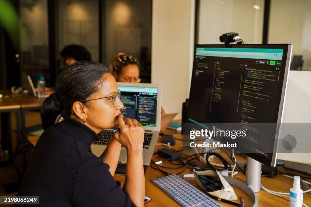 thoughtful female coder looking at desktop pc while sitting in startup office - developer code stock pictures, royalty-free photos & images