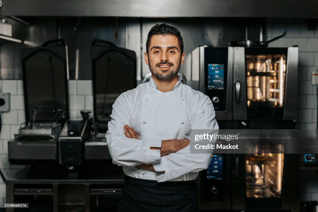 Portrait of confident young male chef wearing white uniform and standing with arms crossed in kitchen at restaurant