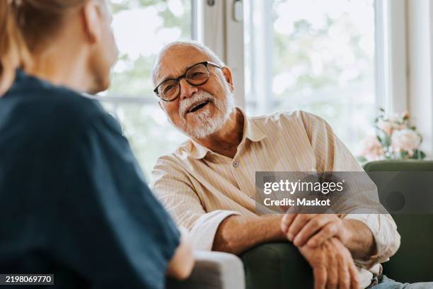 happy senior man laughing while sitting on sofa and talking with female nurse at care home - verouderingsproces stockfoto's en -beelden
