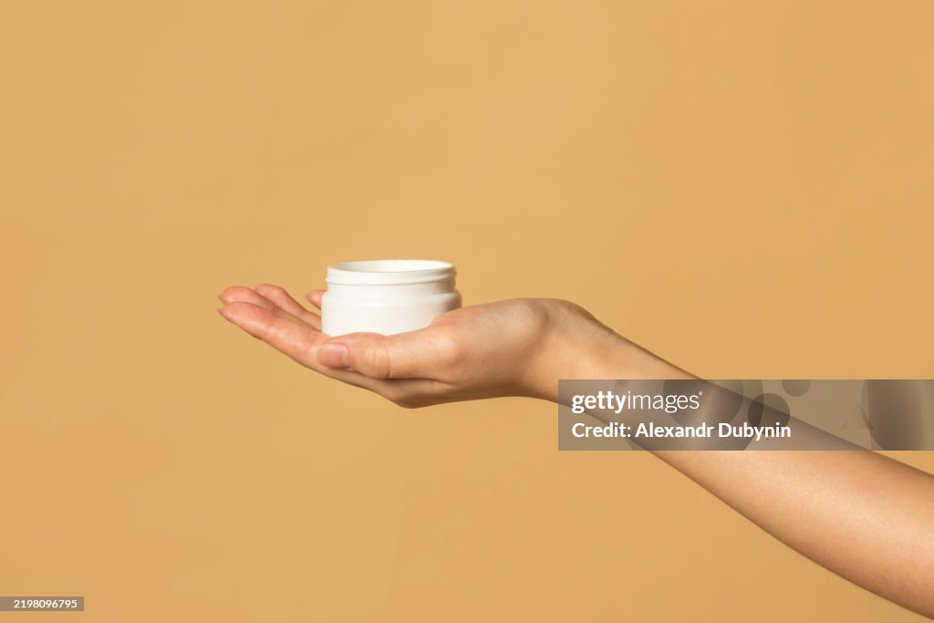 Female hand holding plastic jar with cream on beige background studio shot