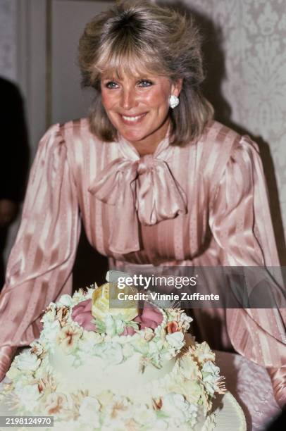American actress Linda Evans standing behind a cake with floral decorations, circa 1985.