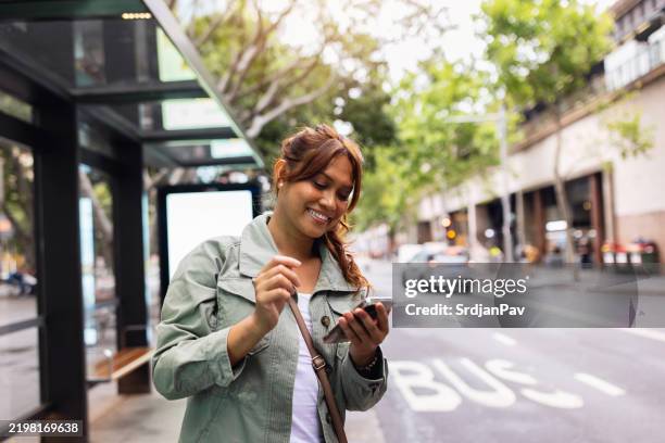 young woman checking smartphone at urban bus stop on bright day - bus stop stock pictures, royalty-free photos & images