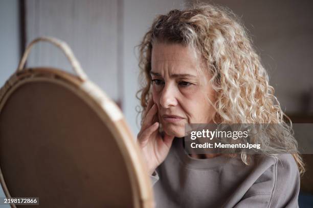 sad middle-aged woman looking in mirror - antirughe foto e immagini stock