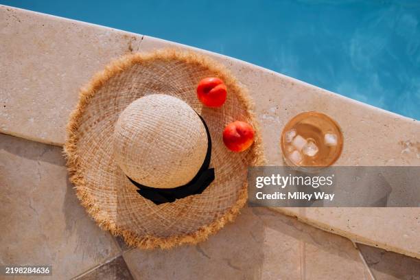 summer relaxation by the pool with straw hat, peaches, and refreshing drink. straw hat and glass of rose wine on blue pool water background. luxury lifestyle and summer vacation content. top view picture. - straw hat stock pictures, royalty-free photos & images