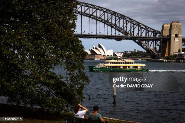 People sit on the foreshore of Sydney Harbour as a ferry passes in front of the Sydney Harbour Bridge and Sydney Opera House during summer on...