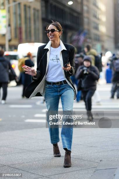 Guest wears black sunglasses, white Calvin Klein Jeans print t-shirt, cream unbuttoned long sleeve shirt, black blazer jacket, black leather belt,...