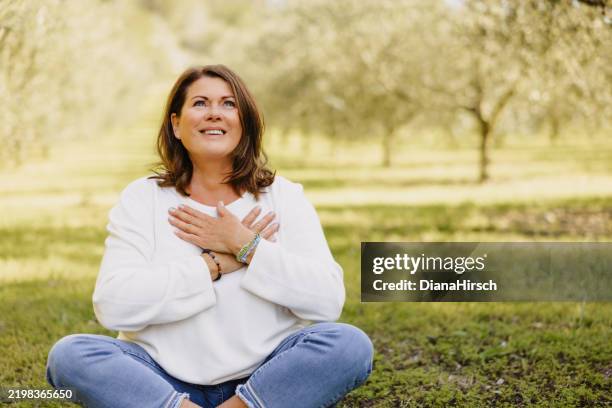 mature woman in her menopausal years sits cross-legged among small and young olive trees with her hands resting gently over her heart in a mindfulness pose - gratidão imagens e fotografias de stock