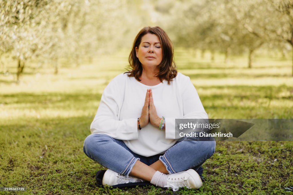 Mature woman in her menopausal years sits cross-legged among young olive trees with her hands gently praying or in a mindfulness pose