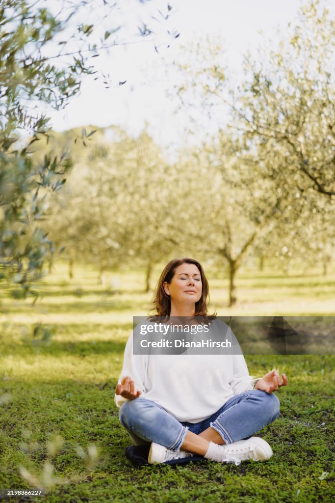 Serene moment as a mature woman in her menopausal years sitting in Padmasana with her hands in Gyan Mudra surrounded by the soft greenery of young olive trees