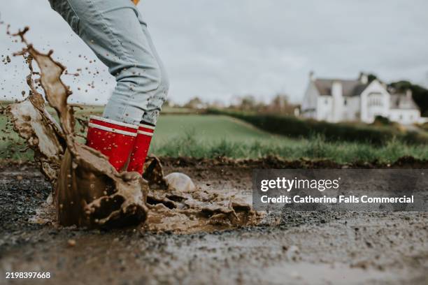 child enjoys splashing in muddy puddle while wearing red rain boots near a green field and farmhouse on a cloudy day - rubberlaars stockfoto's en -beelden
