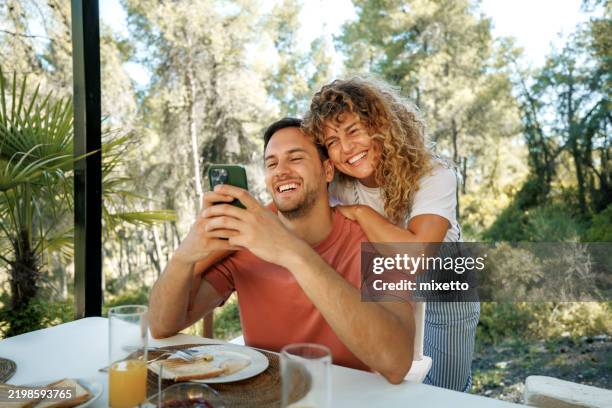 cheerful young couple using smart phone while having breakfast outdoors - couple talking outside stock pictures, royalty-free photos & images