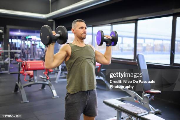 man performing shoulder press with dumbbells in gym - schulterpresse stock-fotos und bilder