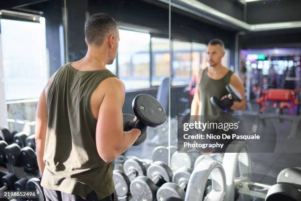 man lifting dumbbells in front of a mirror at the gym - macho stockfoto's en -beelden