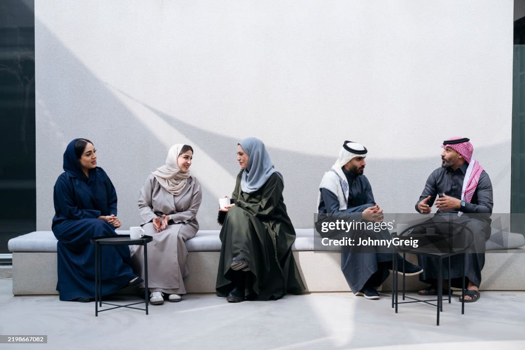 Three businesswomen and two businessmen sitting on bench outside modern office building in traditional Middle Eastern clothing