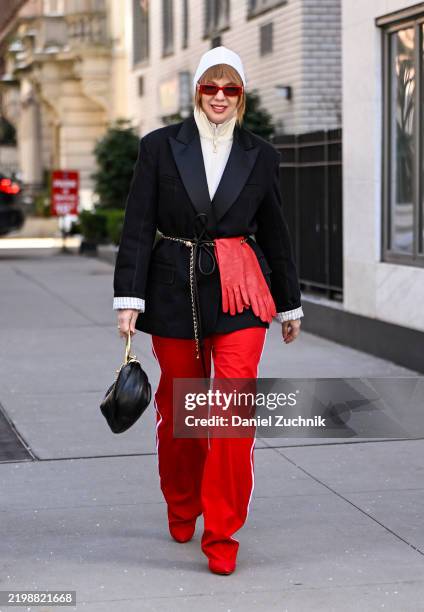 Anna Estrin is seen wearing a black tux jacket, white sweater, red pants, red gloves, gold chain belt, black and gold bag and red sunglasses outside...